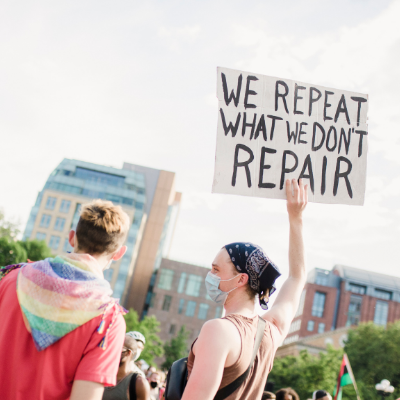 A person holding a sign that reads we repeat what we don't repair at a protest