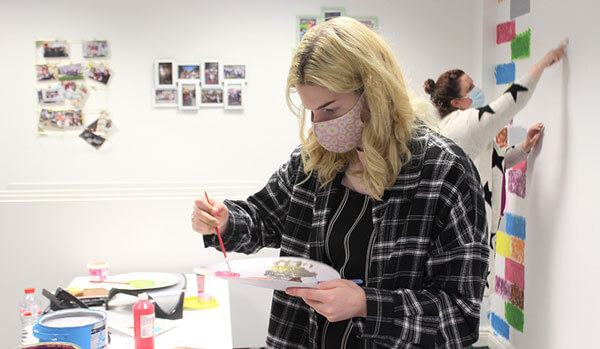 A young woman wearing a mask mixes on a paint palette, with another woman working behind her