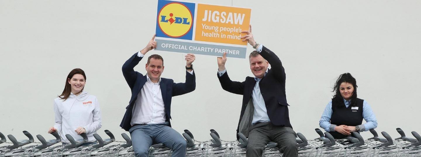 CEO of Lidl and CEO of Jigsaw sit on trolleys while holding up cards with their logos to show their partnership together