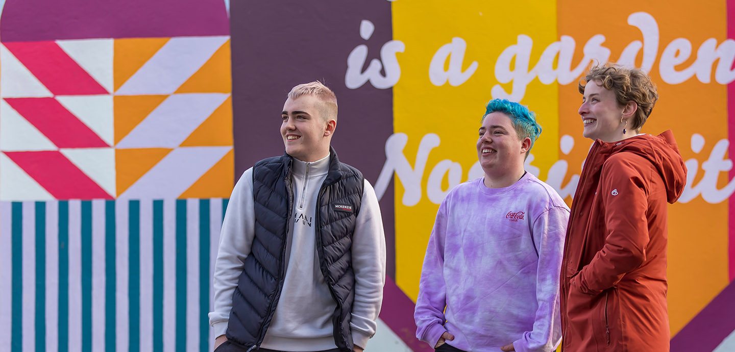 three young people standing smiling in front of a coloured mural