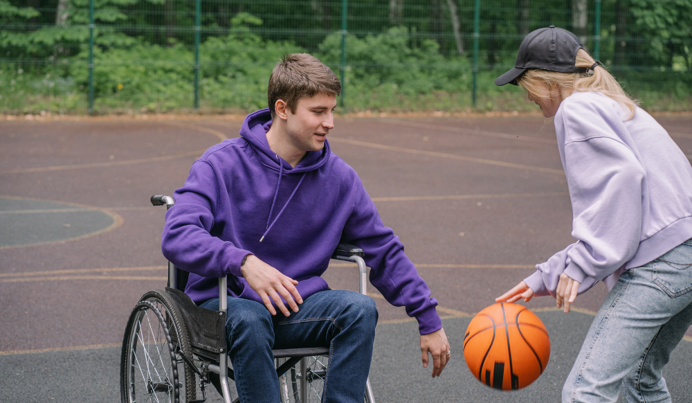 A photograph of a boy and girl playing basketball. The boy is in a wheelchair.
